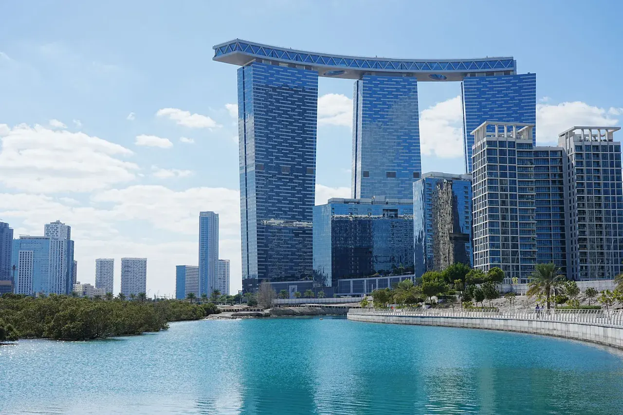 Věže Gate Towers spojené nahoře lávkou na Al Reem Island v Abú Dhabí, před nimi laguna s mangrovy a promenáda u vody